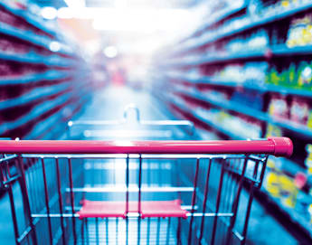 A shopping cart with a red handle is in focus in the foreground of a brightly lit supermarket aisle, surrounded by shelves stocked with a variety of products. The background is blurred, emphasizing the perspective from the shopper's point of view.