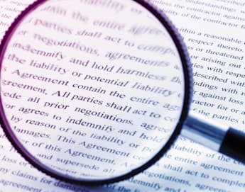 A magnifying glass held over a document with dense text, highlighting words related to agreements, negotiations, and indemnity. The view through the lens is sharp, while the surrounding text is blurred. The image emphasizes the importance of scrutinizing legal documents.