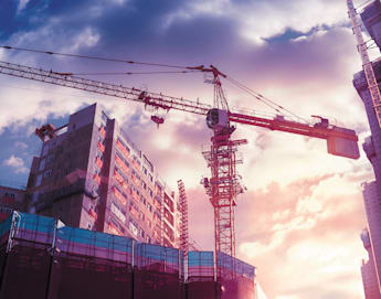 A construction site with tall cranes and buildings under construction. The sky is filled with dramatic clouds illuminated by a colorful sunset, casting a warm glow on the scene. Scaffolding and partially completed structures are prominent in the foreground.