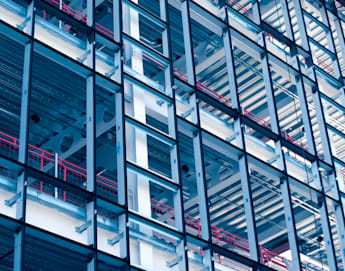 A close-up of a modern building's exterior showcasing its intricate steel framework and glass panels. Red railings and horizontal metallic elements add color and structure to the composition, creating a geometric and industrial aesthetic.