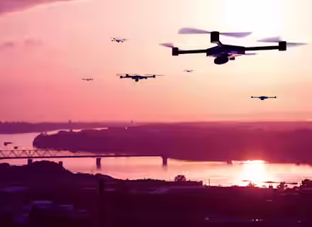Multiple drones are flying over a river at sunset, with a bridge and trees visible in the background. The sky is filled with warm pink and orange hues, reflecting on the water below.