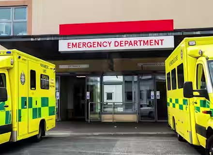 Two yellow ambulances parked outside the entrance to a hospital emergency department, with a large red and white sign above the doorway reading "EMERGENCY DEPARTMENT.