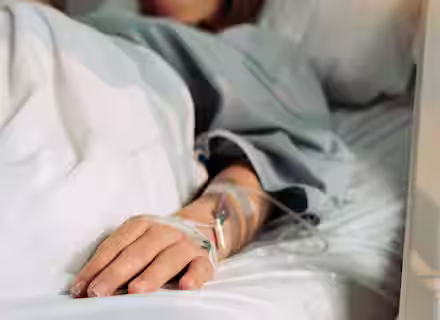 A person lying in a hospital bed with an intravenous (IV) line inserted in their hand, covered by a light blanket, suggesting a medical treatment or hospitalization.