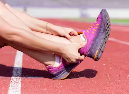 A person sitting on a track holds their ankle in apparent discomfort while wearing purple and orange running shoes.