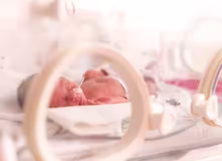 A premature newborn baby lies in an incubator, with a feeding tube taped to their face and surrounded by soft blankets in a neonatal intensive care unit.