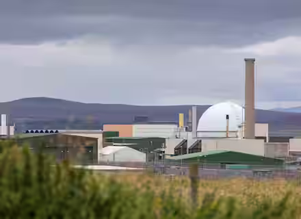 Industrial facility with a large dome-shaped structure and various buildings under a cloudy sky. In the foreground, there are grassy fields and fencing. Hills are visible in the background.