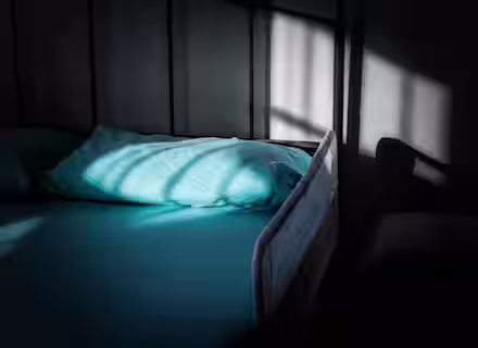 A neatly made bed with a light blue sheet and pillow is partially lit by sunlight streaming through a window, casting shadows. The room appears calm and tranquil.