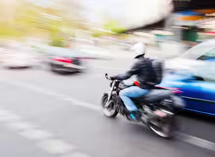 Blurred image of a motorcyclist wearing a helmet and backpack, riding swiftly through city traffic. Surrounding cars appear in motion, creating a dynamic and bustling urban scene.