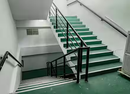 A stairwell with green steps and black railings. The stairway is split halfway with a landing, leading to a lower flight of stairs. The walls are painted white, and there is a vent on the wall above the landing. The area is well-lit with fluorescent lighting.