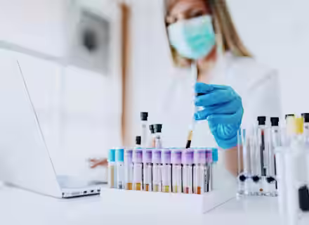 A lab technician wearing a mask and blue gloves handles a test tube with blood samples, working at a desk with a laptop and racks of test tubes in a bright laboratory setting.