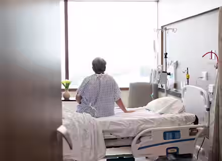 A person with short gray hair wearing a hospital gown sits on the edge of a hospital bed, looking out a large window. The room is equipped with medical equipment, including an IV stand and monitor, and a potted plant is placed on a table by the window.
