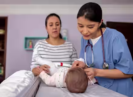 A nurse examines a baby lying on an exam table while the mother stands nearby, watching attentively in a medical office.
