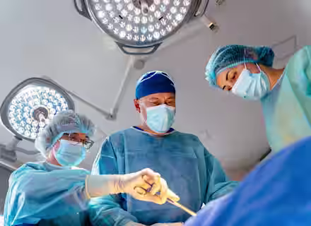 Three surgeons in blue scrubs, masks, and hair covers perform surgery under bright operating room lights, focusing intently on their work.