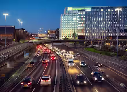 Evening scene of a busy urban highway with multiple lanes of vehicles under an illuminated overpass. A modern office building with bright lights stands prominently in the background against the twilight sky. Several vehicles have their headlights on.