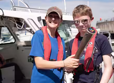 Two people wearing red life jackets and sunglasses smile on a boat. One holds a metal object while the other places a hand on their shoulder. The background shows a sunny marina with boats and a pier.