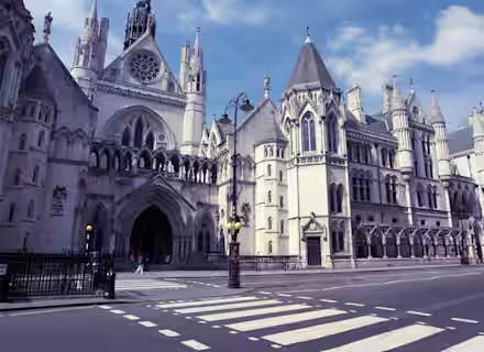 A wide view of the exterior of the Royal Courts of Justice in London on a clear day. The Gothic-style building features pointed arches, intricate stonework, and large windows. The street in front has a zebra crossing and few pedestrians.