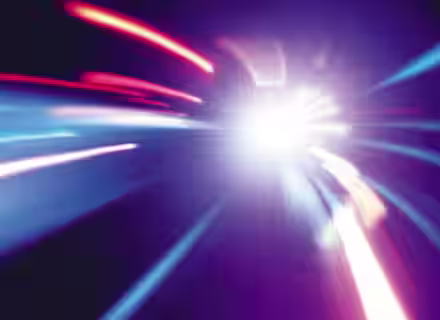 A dynamic, abstract image with streaks of light in blue, red, and purple hues radiating from a bright, central point. The lights create a sense of motion and speed, with blurred lines and a gradient background adding to the energetic effect.