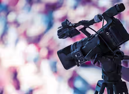 A professional video camera on a tripod is focused on an event. The background is blurred with colorful, out-of-focus shapes, suggesting a vibrant setting with a crowd or audience. The camera is equipped with various attachments and appears ready for filming.