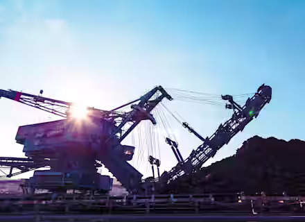 A large industrial excavator machine is working at a mining site. The sun shines brightly in the background, casting a glow on the machinery. Piles of excavated earth and rocks are visible beside the machine under a clear blue sky.