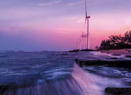 A serene sunset over a coastal area with wind turbines in the distance. The sky is painted in shades of pink, purple, and blue. Waves are crashing against a rocky shore, creating a dynamic foreground, while trees line the horizon.