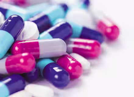 A close-up image of a pile of various pills and capsules. The capsules are predominantly in shades of blue, pink, white, and purple, while the pills are mostly white. The background is a plain, light surface.