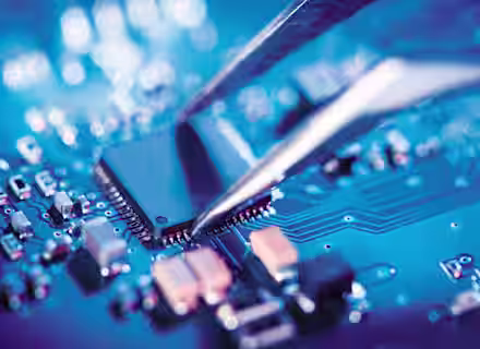 Close-up of tweezers placing a microchip onto a blue printed circuit board (PCB), showcasing electronic components and intricate conductive pathways, with a blurred background creating a focused detail on the microchip and its precise positioning.