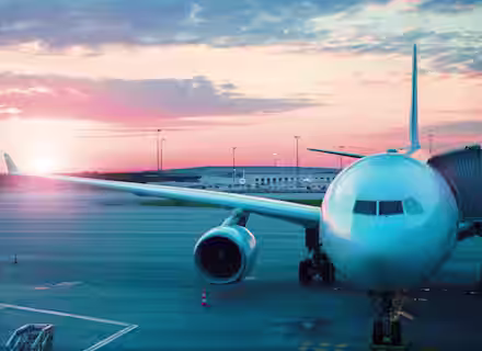 A large passenger airplane is parked at an airport gate during sunset. The sky features a mix of pink, orange, and blue hues while the sun is partially visible near the horizon. The airplane is connected to the gate by a jet bridge.