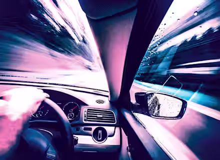 A blurred, high-speed view from inside a car, focusing on the driver's hand on the steering wheel, dashboard, and side mirror. The outside scenery and road ahead are motion-blurred, giving a sense of rapid movement and speed.