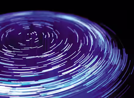 A circular, abstract light pattern with bright blue and purple streaks radiating outward on a dark background, resembling long-exposure photography of spinning lights. The streaks create a sense of motion and depth, forming a visually dynamic composition.
