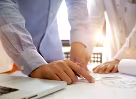Two people wearing light blue shirts are reviewing architectural blueprints on a table in a brightly lit room. One person is pointing at the blueprints with a pen, while the other rests their hands on the table. A laptop is visible to one side.