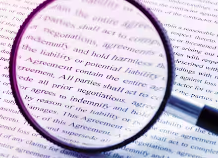 A magnifying glass lying on top of a document, enlarging the text beneath it. The document contains formal legal language and is printed in black font on white paper. The magnified text mentions liability and agreements.