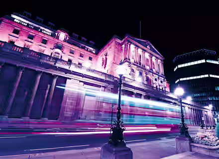 A historic building illuminated with pink and purple lights at night. Long exposure creates light trails from passing vehicles, enhancing the dynamic scene. Street lamps and modern buildings in the background contrast with the classical architecture.