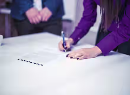 A person in a purple shirt signs a document labeled "CONTRACT" on a white table. Another person stands in the background with their hands clasped. A small white cup is to the left of the contract on the table.
