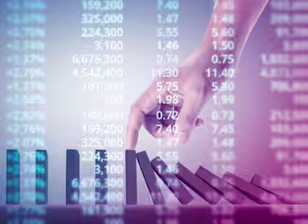 A hand is seen stopping a row of falling dominoes against a backdrop of fluctuating stock market numbers. The image symbolizes intervention and stability amidst financial fluctuations.