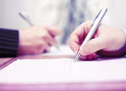 Close-up of two people writing on documents with silver pens. The focus is on their hands, one person wearing a suit jacket and the other in a light blue shirt, both engaged in what appears to be a formal signing or writing activity.