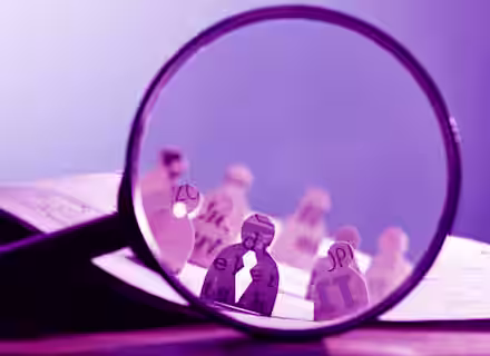 A large magnifying glass focuses on paper cut-outs of human figures, each decorated with various letters and text passages. The background shows more paper figures out of focus, positioned on an open book, creating a scene with text and paper characters.