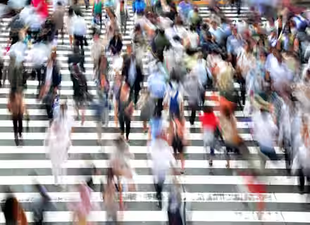 A blurred image of numerous people walking across a large, busy, zebra-striped crosswalk. The motion blur captures the hustle and bustle of the crowd, giving a sense of fast-paced urban life. The scene is vibrant with diverse clothing colors and patterns.