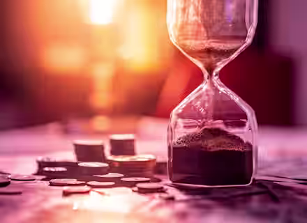 A close-up of an hourglass with sand flowing into the lower chamber, placed on a table scattered with coins and a few stacks of coins. The background is blurred with a warm, glowing light, creating a soft and nostalgic ambiance.