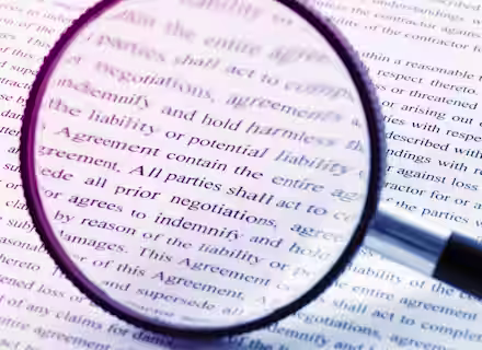 A magnifying glass held over a document with dense text, highlighting words related to agreements, negotiations, and indemnity. The view through the lens is sharp, while the surrounding text is blurred. The image emphasizes the importance of scrutinizing legal documents.