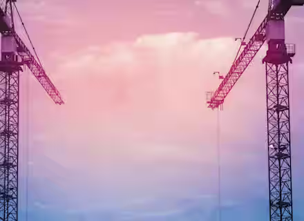 Two tall construction cranes are silhouetted against a vibrant, multi-colored sky with hues of pink, purple, and blue. The cranes face each other, forming a symmetrical frame, while wispy clouds drift in the background.