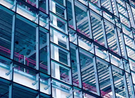 A close-up of a modern building's exterior showcasing its intricate steel framework and glass panels. Red railings and horizontal metallic elements add color and structure to the composition, creating a geometric and industrial aesthetic.