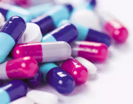 A close-up image of a pile of various pills and capsules. The capsules are predominantly in shades of blue, pink, white, and purple, while the pills are mostly white. The background is a plain, light surface.