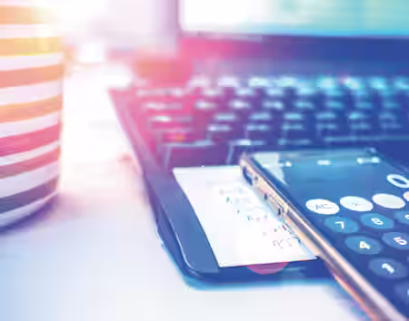 A close-up of a desk workspace with a striped mug, a computer keyboard, and a smartphone displaying a calculator app. A piece of paper with handwritten notes is beneath the phone. Sunlight creates lens flare effects on the image.