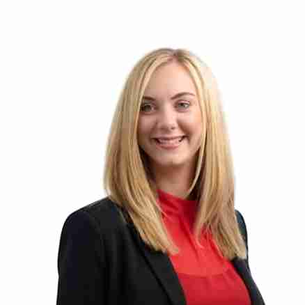 A woman with long blonde hair is smiling and wearing a black blazer over a red top. The background is white.