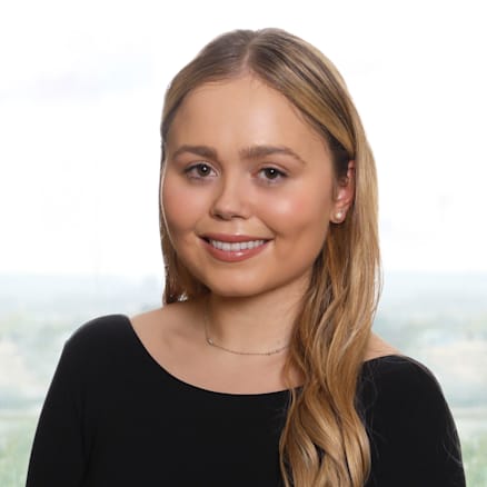 A young woman with long blonde hair smiles at the camera. She is wearing a black, long-sleeved top and a subtle necklace. The background is light and appears slightly blurred.