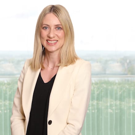 A woman with straight blonde hair, wearing a black top and white blazer, smiles while standing in front of a large window overlooking a cityscape.