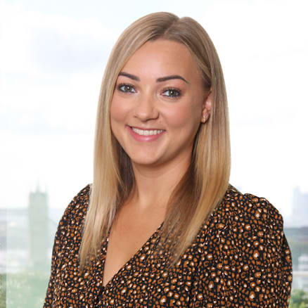 A woman with long, straight blonde hair is smiling at the camera. She is wearing a black dress with a small orange and white pattern. The blurred background indicates she is indoors with a window showing a cityscape behind her.