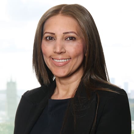 A woman with long, straight brown hair is smiling at the camera. She is wearing a dark blazer over a matching top. The background is blurred, showing what appears to be a cityscape with buildings and greenery.