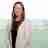 A woman with long brown hair wearing a light gray blazer and a black top smiles at the camera. She stands against a blurry outdoor background featuring green foliage and a hazy skyline.