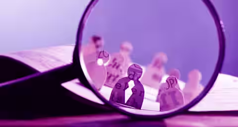 A large magnifying glass focuses on paper cut-outs of human figures, each decorated with various letters and text passages. The background shows more paper figures out of focus, positioned on an open book, creating a scene with text and paper characters.